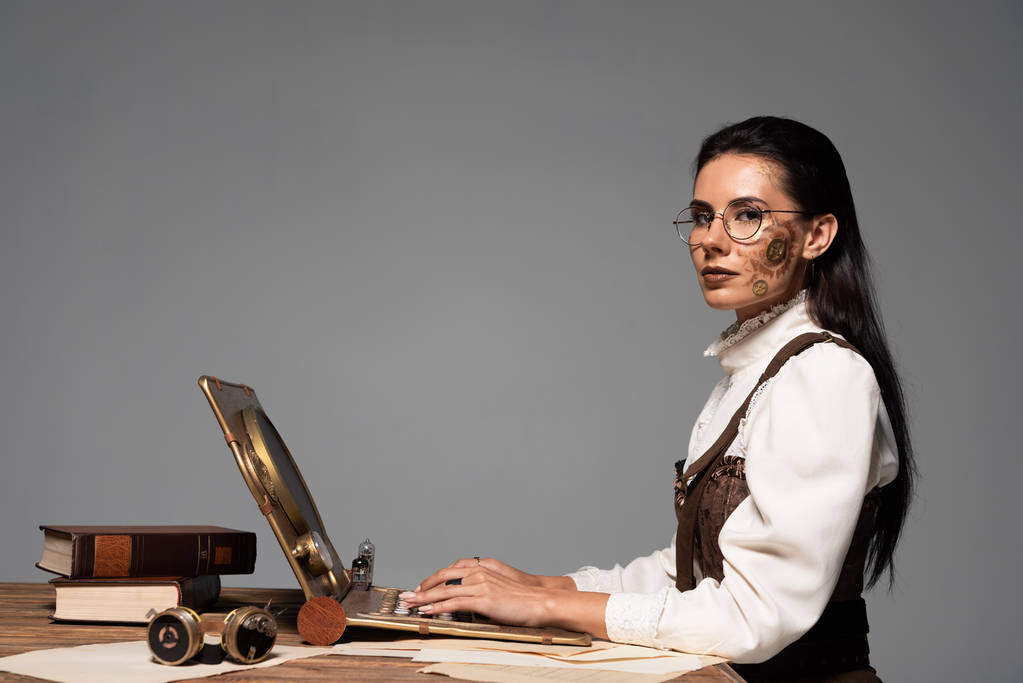 привлекательная стимпанк-женщина с ноутбуком на рабочем месте, изолированная от серых
 - Фото, изображение