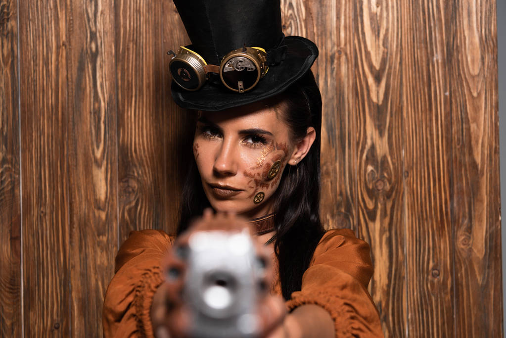selective focus of serious steampunk woman in top hat with goggles aiming with pistol at camera on wooden - Photo, Image
