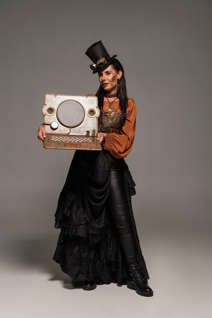 full length view of woman in top hat with goggles showing steampunk laptop on grey - Photo, Image