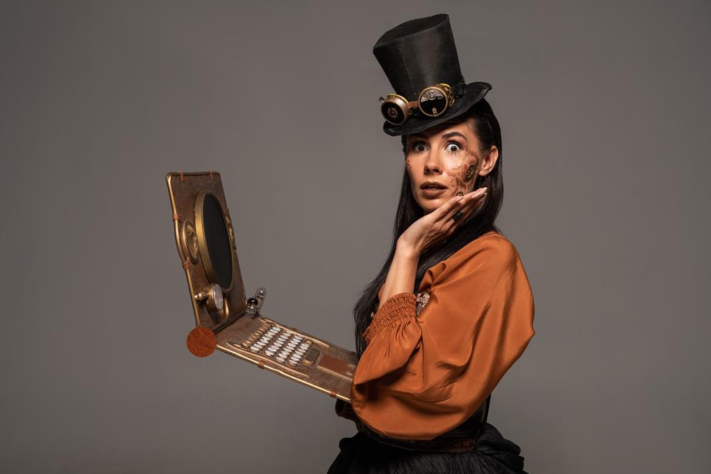 mulher chocada no chapéu superior com óculos usando laptop steampunk isolado em cinza
 - Foto, Imagem