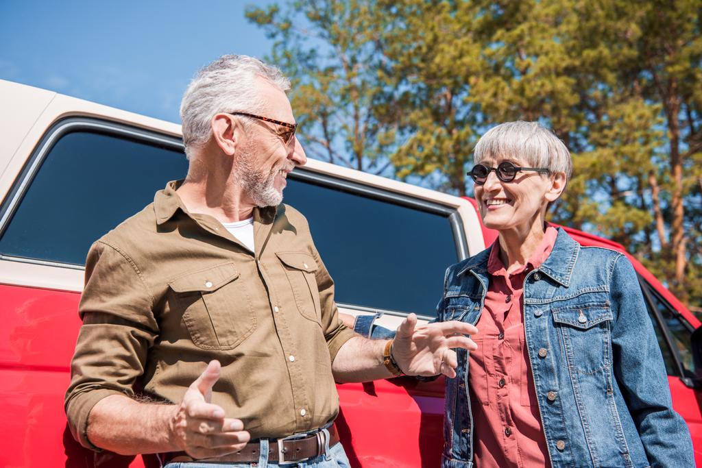 Seniorenpaar mit Sonnenbrille steht neben rotem Auto und schaut sich an sonnigem Tag an - Foto, Bild