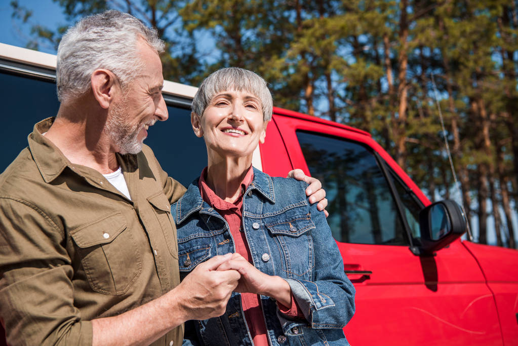 χαμογελαστοί ηλικιωμένοι τουρίστες στέκονται κοντά στο κόκκινο αυτοκίνητο, κρατώντας τα χέρια και αγκαλιάζοντας την ηλιόλουστη μέρα - Φωτογραφία, εικόνα
