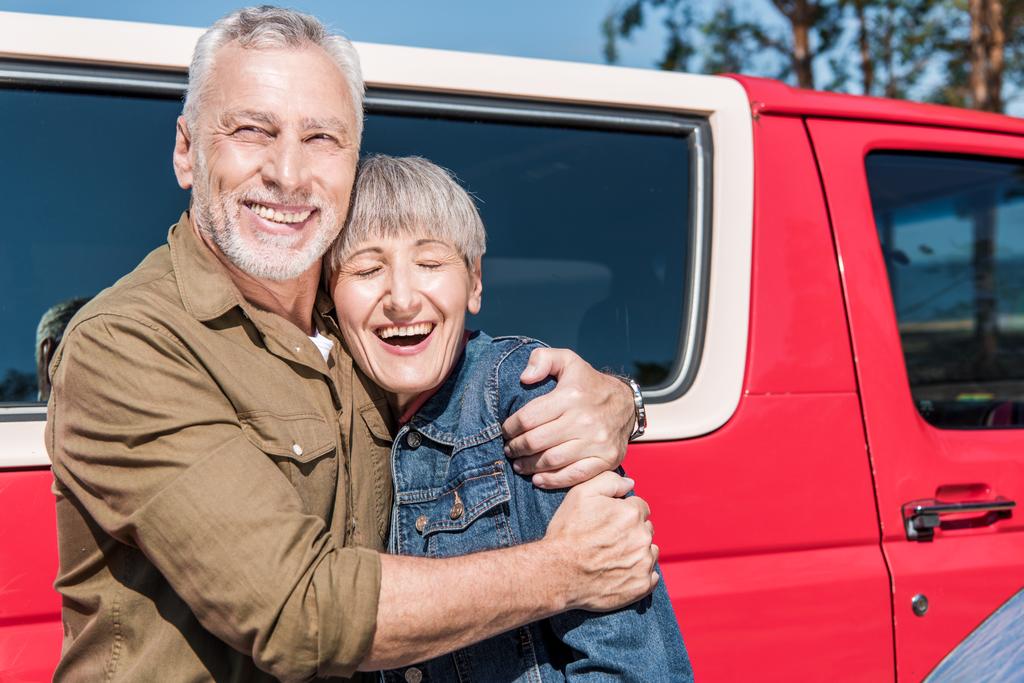 χαμογελαστοί ηλικιωμένοι τουρίστες που στέκονται κοντά στο κόκκινο αυτοκίνητο και αγκαλιάζονται την ηλιόλουστη μέρα - Φωτογραφία, εικόνα