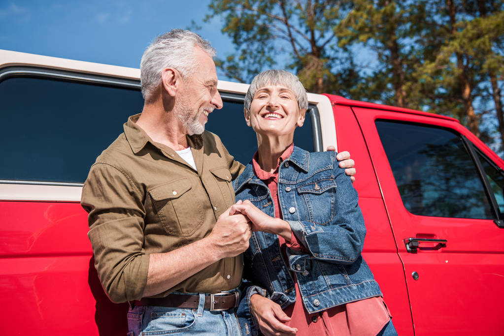 sorridente coppia di turisti anziani in piedi vicino alla macchina rossa, tenendosi per mano e abbracciando nella giornata di sole
 - Foto, immagini