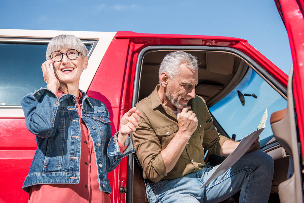 mujer mayor en gafas hablando en teléfono inteligente, mientras que el marido sentado en el coche y mirando el mapa
 - Foto, Imagen