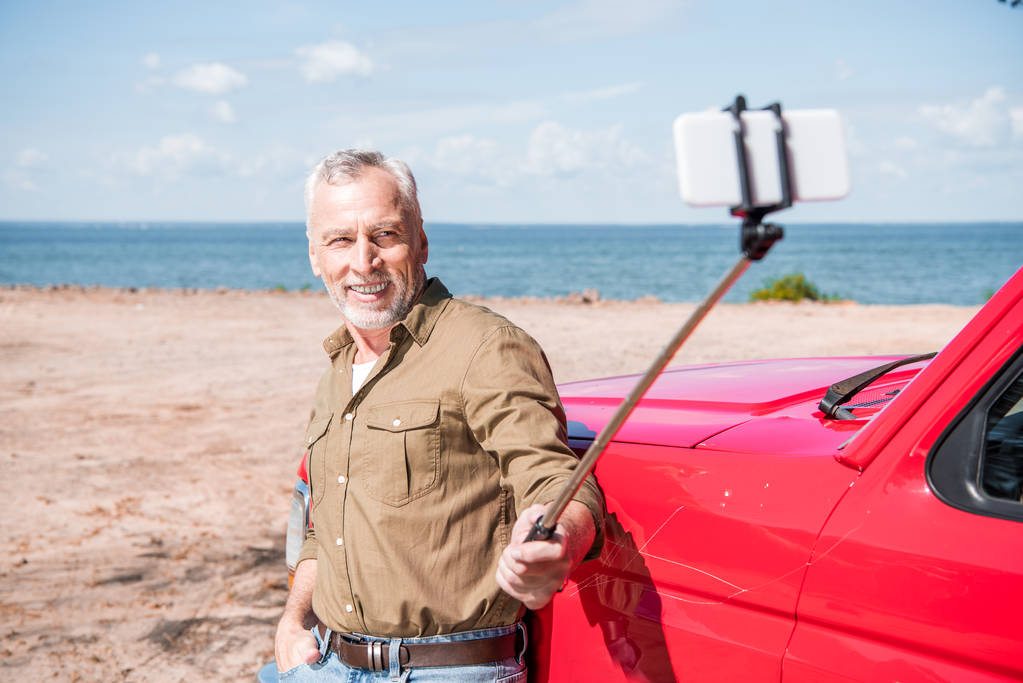uśmiechnięty starszy człowiek stoi w pobliżu czerwonego samochodu i biorąc selfie w słoneczny dzień - Zdjęcie, obraz