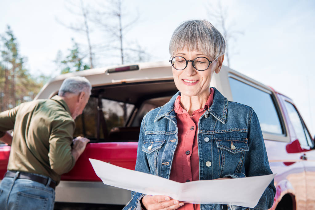 Lächelnde Seniorin mit Karte, während Mann an sonnigem Tag neben Auto steht - Foto, Bild