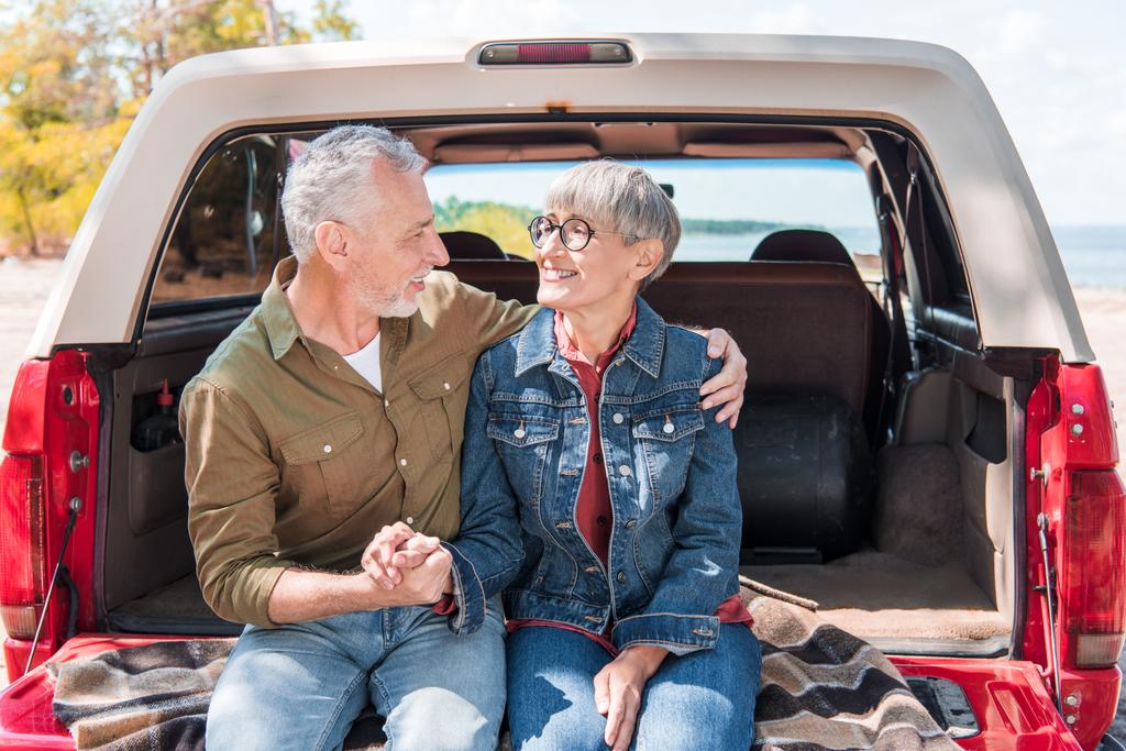 Lächelndes Senioren-Paar umarmt sich und blickt sich in der Nähe des Autos an - Foto, Bild
