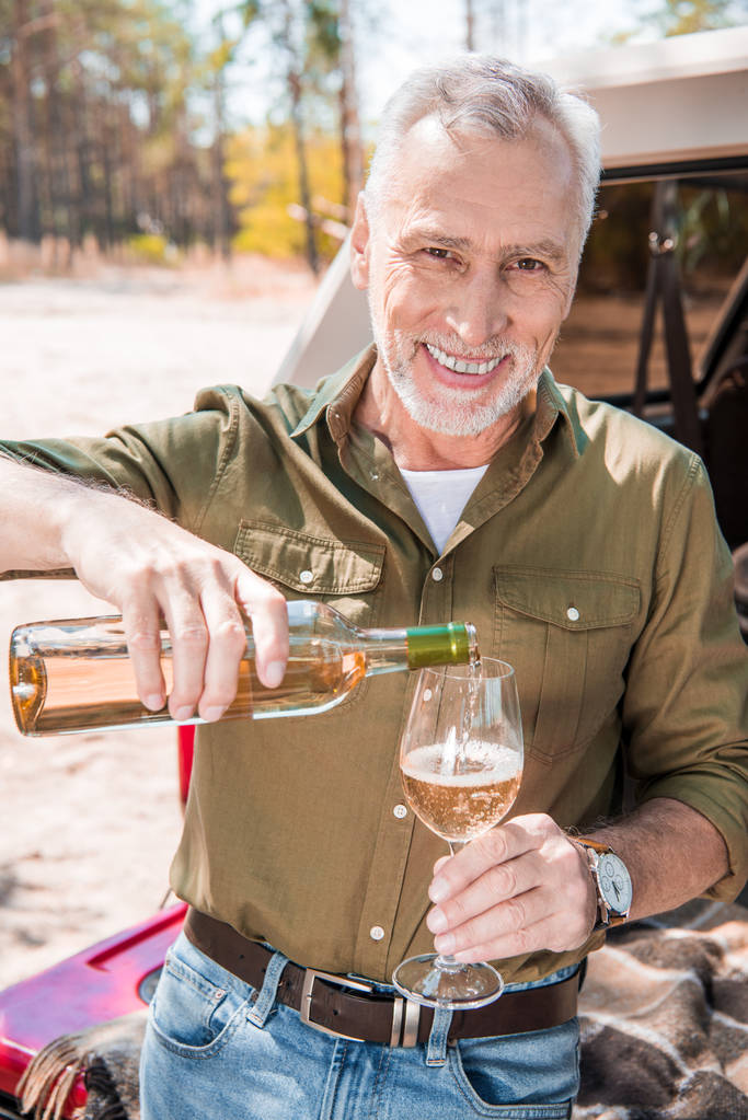smiling senior man standing near car and pouring wine in wine glass - Photo, Image