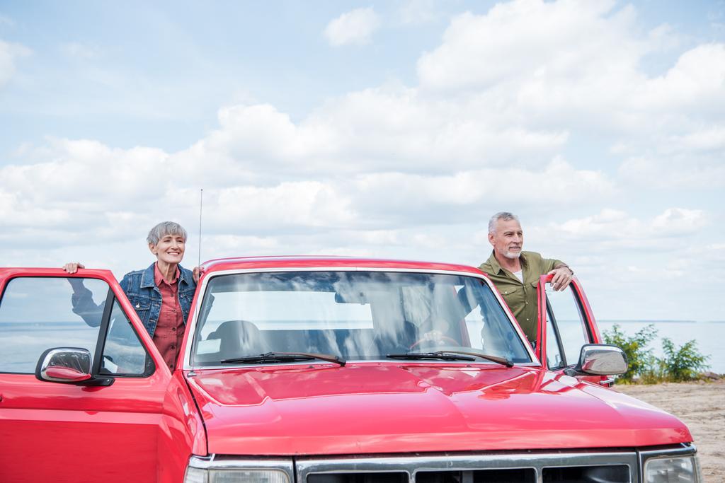 γηραιότερος ζευγάρι τουριστών που στέκονται κοντά στο κόκκινο αυτοκίνητο στην παραλία - Φωτογραφία, εικόνα