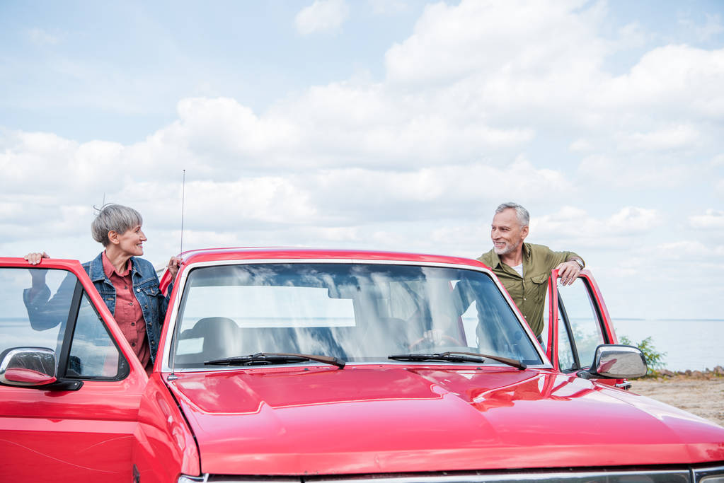 улыбающаяся пожилая пара туристов, стоящих рядом с красной машиной и смотрящих друг на друга на пляже
 - Фото, изображение