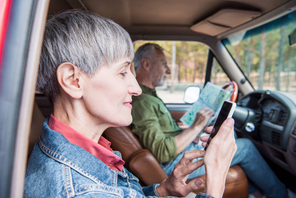 Seitenansicht einer lächelnden Seniorin mit Smartphone mit leerem Bildschirm, während ihr Mann im Auto auf die Landkarte schaut - Foto, Bild