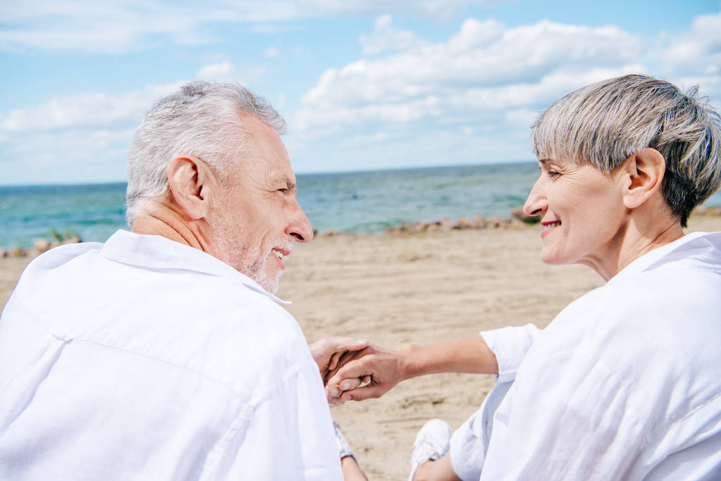 hymyilevä vanhempi pari kädestä pitäen ja katsellen toisiaan rannalla
 - Valokuva, kuva