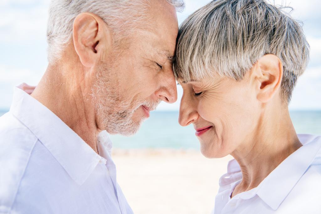 πλευρική όψη του χαμογελαστή ζευγάρι ηλικιωμένων αγγίζοντας τα μέτωπα με κλειστά μάτια στην παραλία - Φωτογραφία, εικόνα