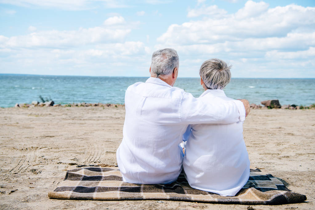 vista trasera de pareja mayor en camisas blancas sentada en manta y abrazándose en la playa
 - Foto, imagen