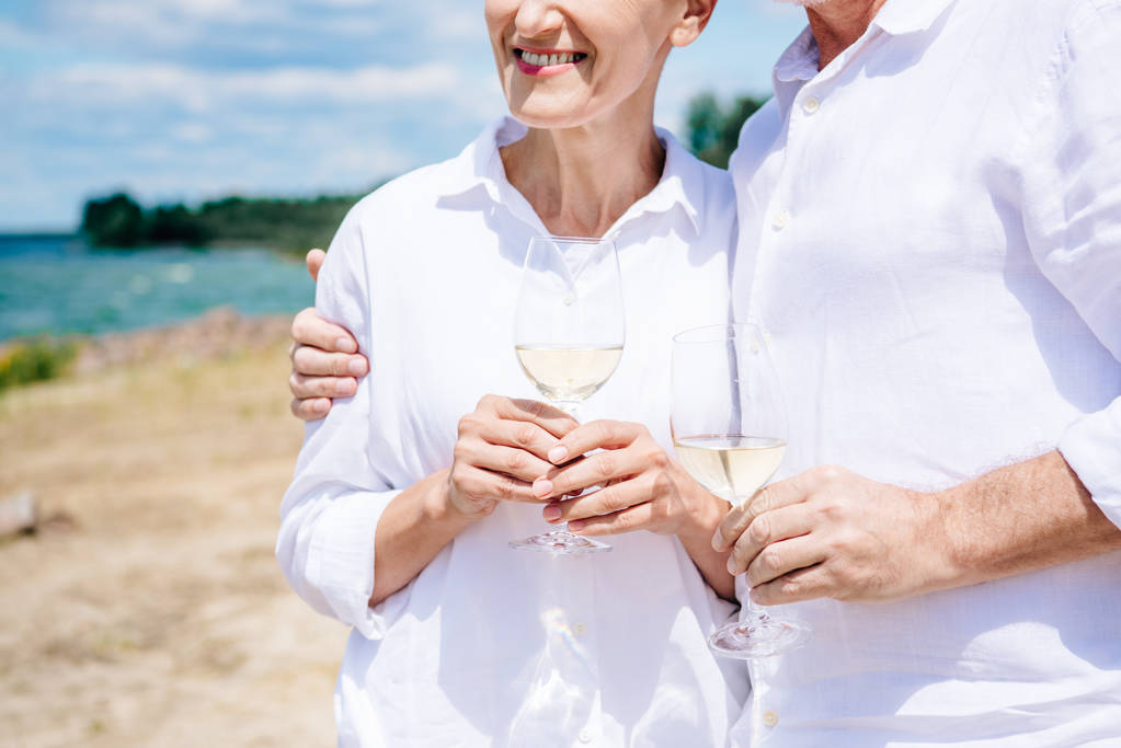 περικομμένη θέα του χαμογελαστή ζευγάρι ηλικιωμένων Αγκαλιάζοντας και κρατώντας ποτήρια κρασιού με κρασί στην παραλία - Φωτογραφία, εικόνα