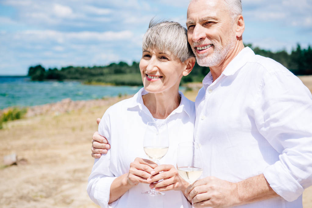 uśmiechnięta Starsza para obejmując i trzymając kieliszki do wina z winem na plaży - Zdjęcie, obraz