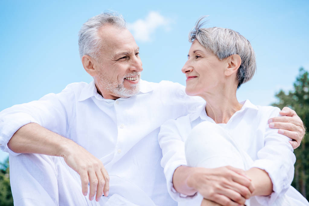 счастливая улыбающаяся пожилая пара в белых рубашках обнимающая и смотрящая друг на друга под голубым небом
 - Фото, изображение