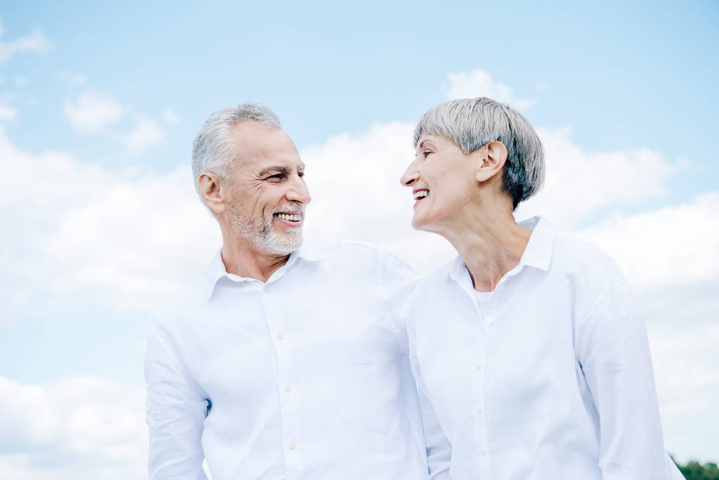 onnellinen hymyilevä vanhempi pari valkoisissa paidoissa katsomassa toisiaan sinisen taivaan alla
 - Valokuva, kuva