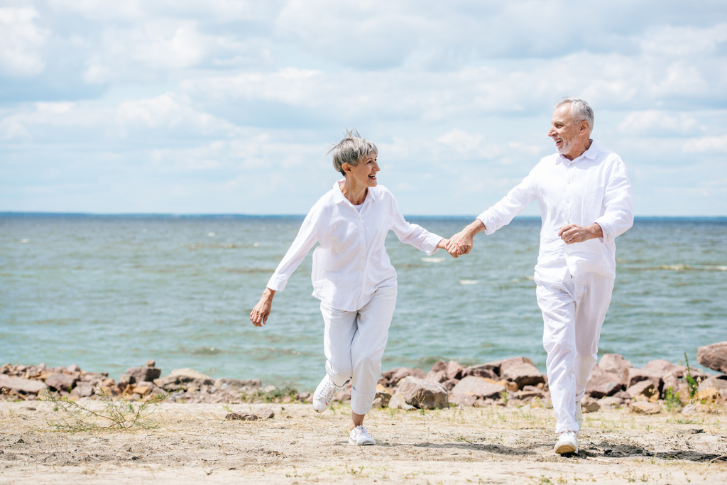 vista completa de la pareja de ancianos tomados de la mano y mirándose en la playa
 - Foto, Imagen