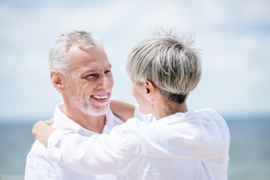 счастливая пожилая пара в белых рубашках обнимающая и смотрящая друг на друга под голубым небом
 - Фото, изображение