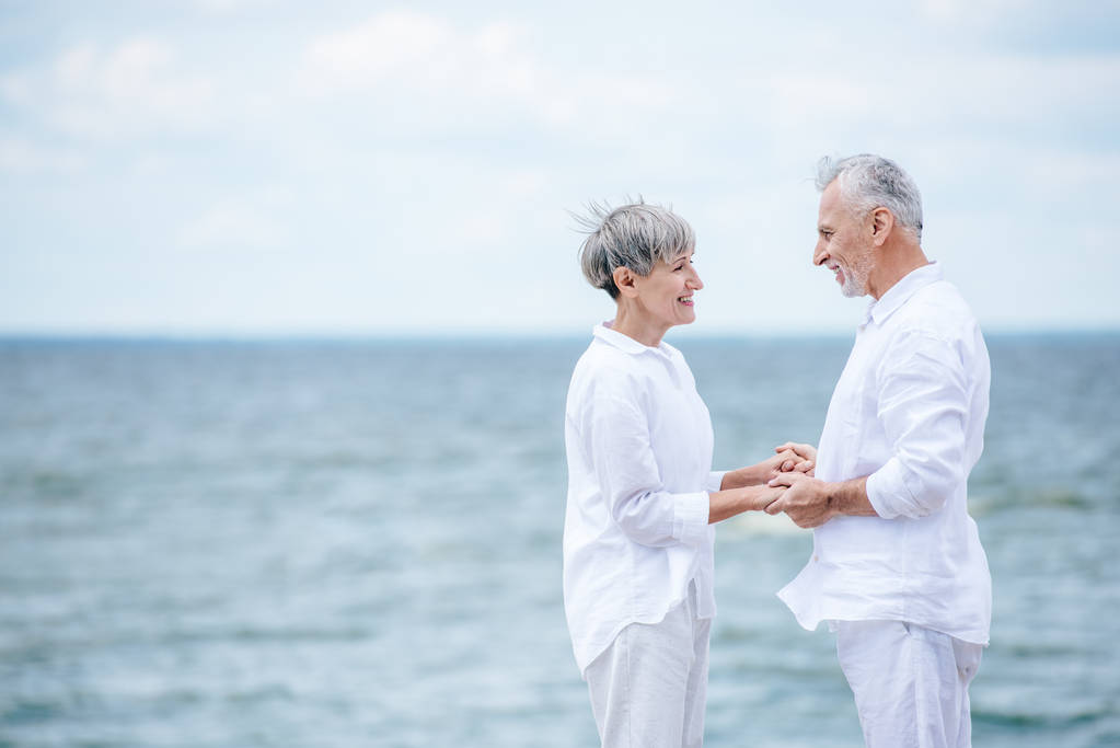 pohled ze strany šťastného staršího páru, který se drží za ruce a dívá se na sebe poblíž řeky - Fotografie, Obrázek