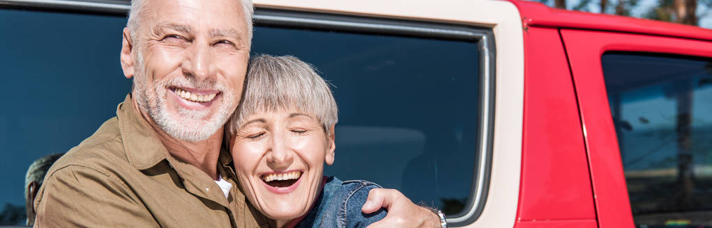 Panoramablick auf ein glückliches älteres Touristenpaar, das neben dem Auto steht und sich lächelnd umarmt - Foto, Bild
