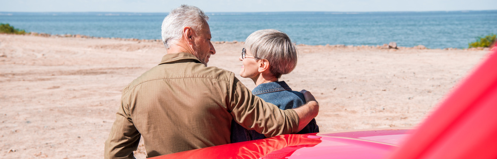 Panoramablick auf ein älteres Touristenpaar, das neben dem Auto steht und sich am Strand umarmt - Foto, Bild