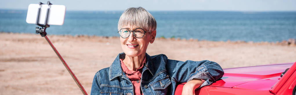 vista panoramica della donna anziana sorridente in piedi vicino alla macchina e scattare selfie in spiaggia
 - Foto, immagini