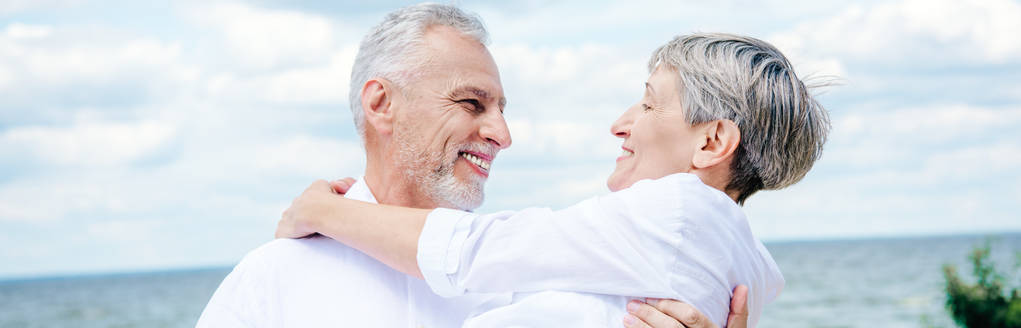 Panoramablick auf einen lächelnden Senior, der seine Frau unter blauem Himmel abhebt - Foto, Bild
