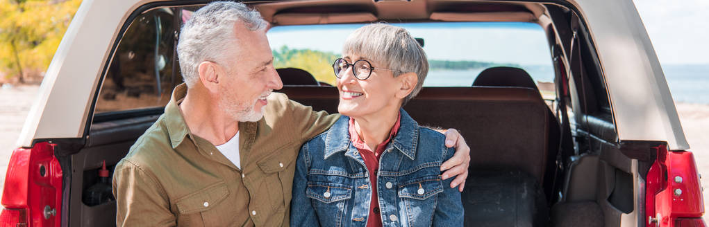panoramic view of smiling senior couple embracing and looking at each other near car - Photo, Image