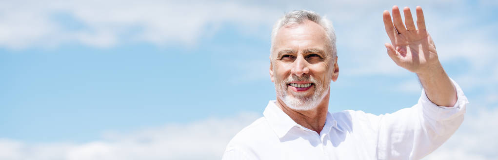 panoraamanäkymä hymyilevä vanhempi mies valkoisessa paidassa heiluttaa käsi sinisen taivaan alla
 - Valokuva, kuva