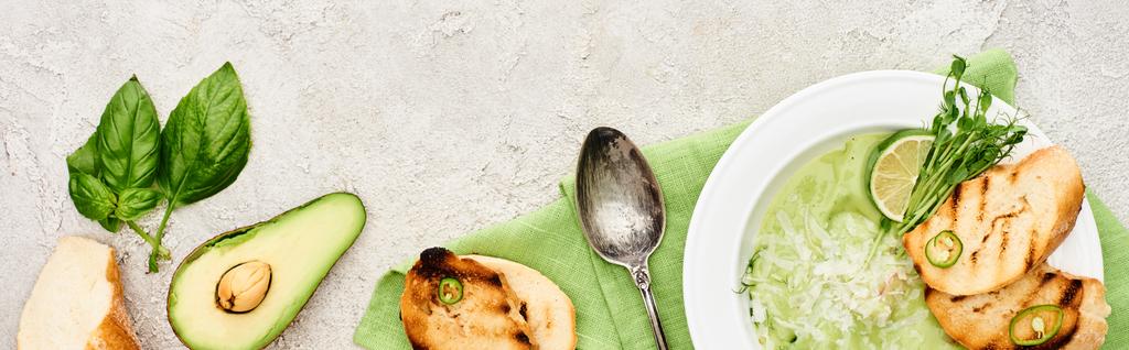панорамный снимок вкусного сливочно-зеленого овощного супа с гренками подается с ложкой на салфетке рядом со свежими ингредиентами
 - Фото, изображение