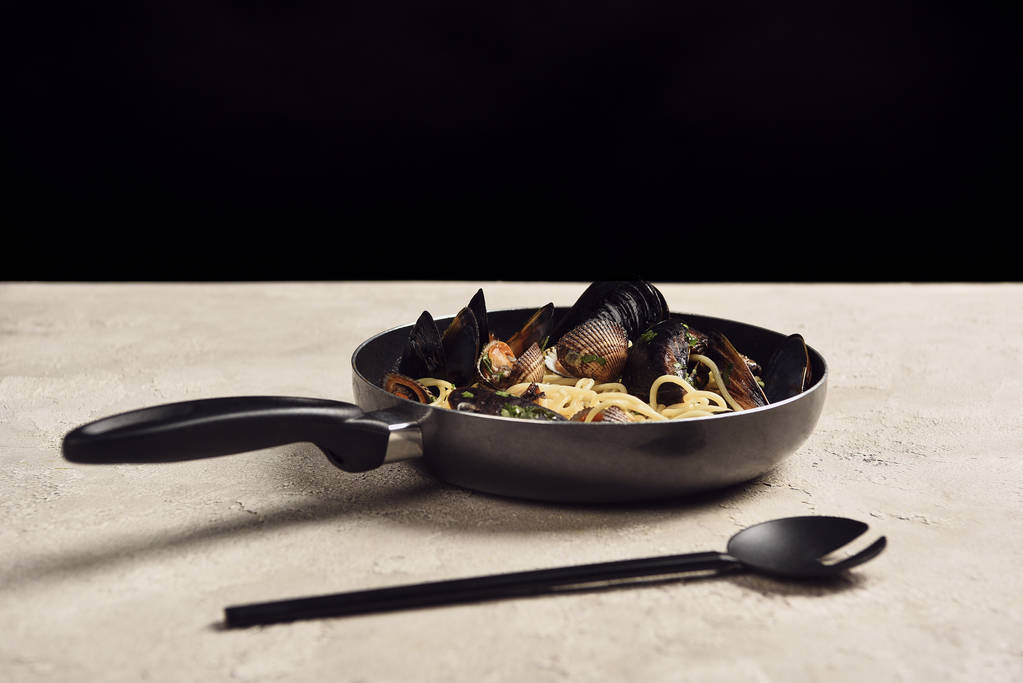 deliciosa pasta italiana con mariscos servida en sartén cerca de espátula aislada en negro
 - Foto, Imagen