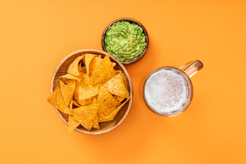 top view of crispy nachos in wooden bowl near guacamole and beer on orange background, Mexican cuisine - Photo, Image
