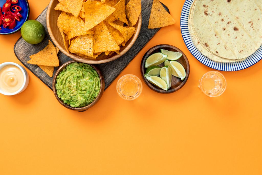 Mexican nachos served on wooden cutting board with guacamole, cheese sauce, peppers, limes, Tequila and tortillas on orange background with copy space - Photo, Image
