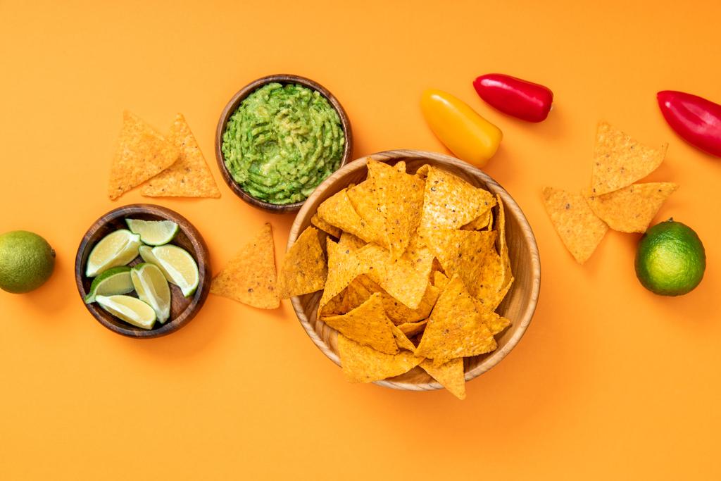 Top uitzicht van knapperige Mexicaanse nacho's, guacamole, paprika en limotjes op oranje achtergrond - Foto, afbeelding