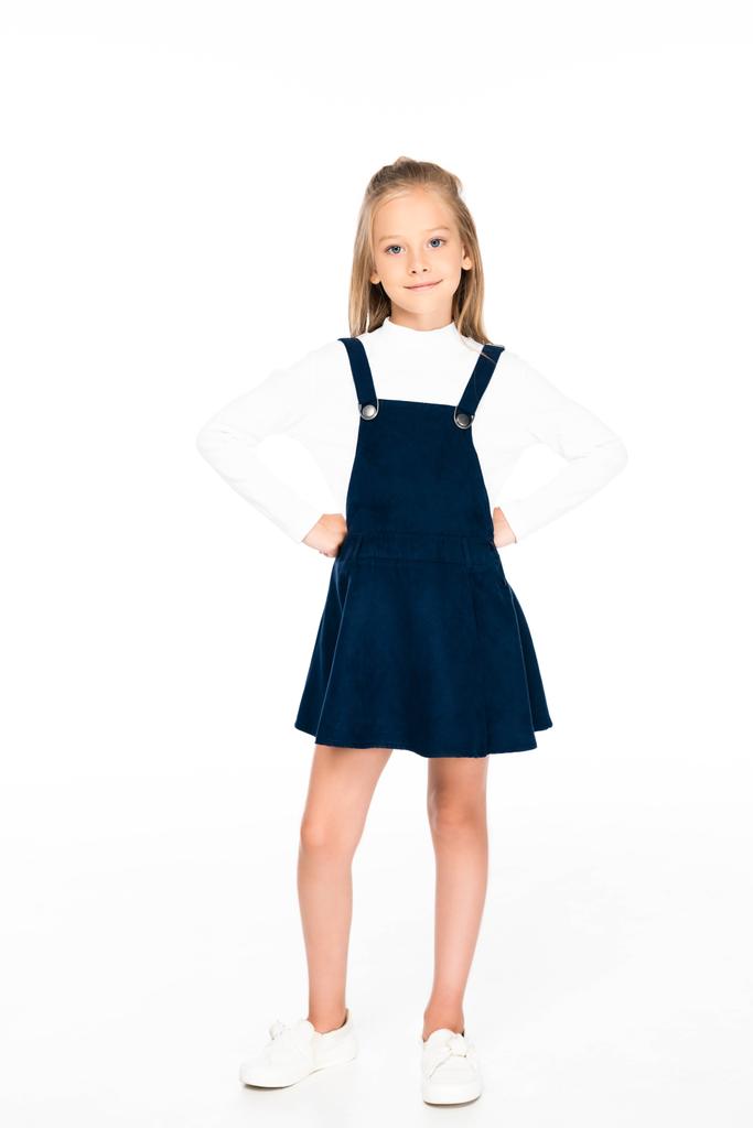 full length view of cute schoolgirl holding hands on hips while posing at camera on white background - Photo, Image