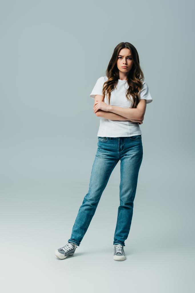 грустная красивая девушка в белой футболке и джинсах со скрещенными руками, изолированными на сером
 - Фото, изображение