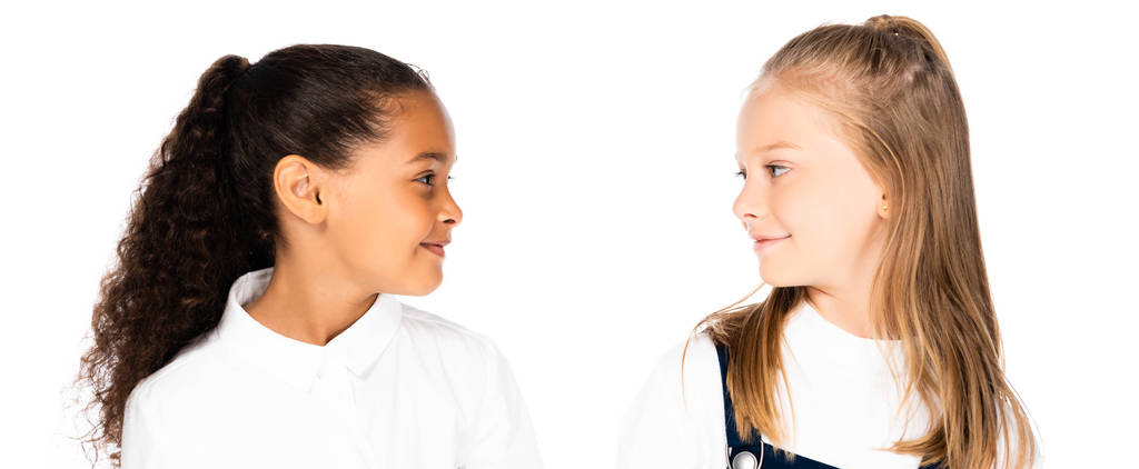 панорамный снимок двух улыбающихся мультикультурных школьниц, смотрящих друг на друга изолированно на белом
 - Фото, изображение