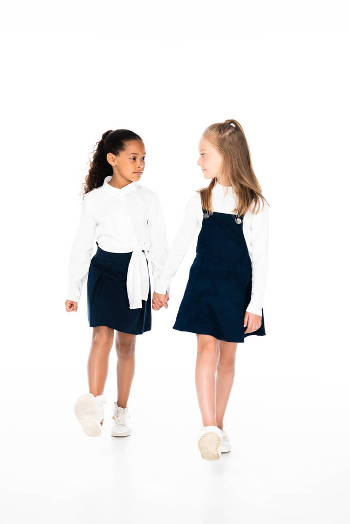 volledige lengte uitzicht van twee multiculturele schoolmeisjes houden van handen en lopen samen op witte achtergrond - Foto, afbeelding