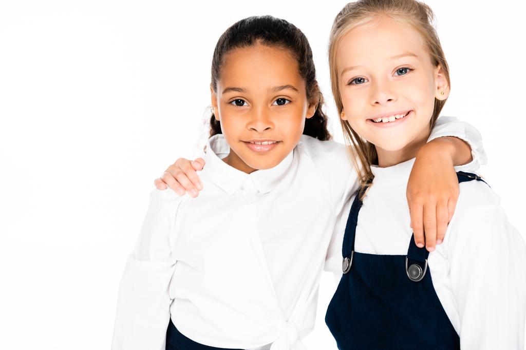 two happy multicultural schoolgirls hugging and smiling at camera isolated on white - Photo, Image