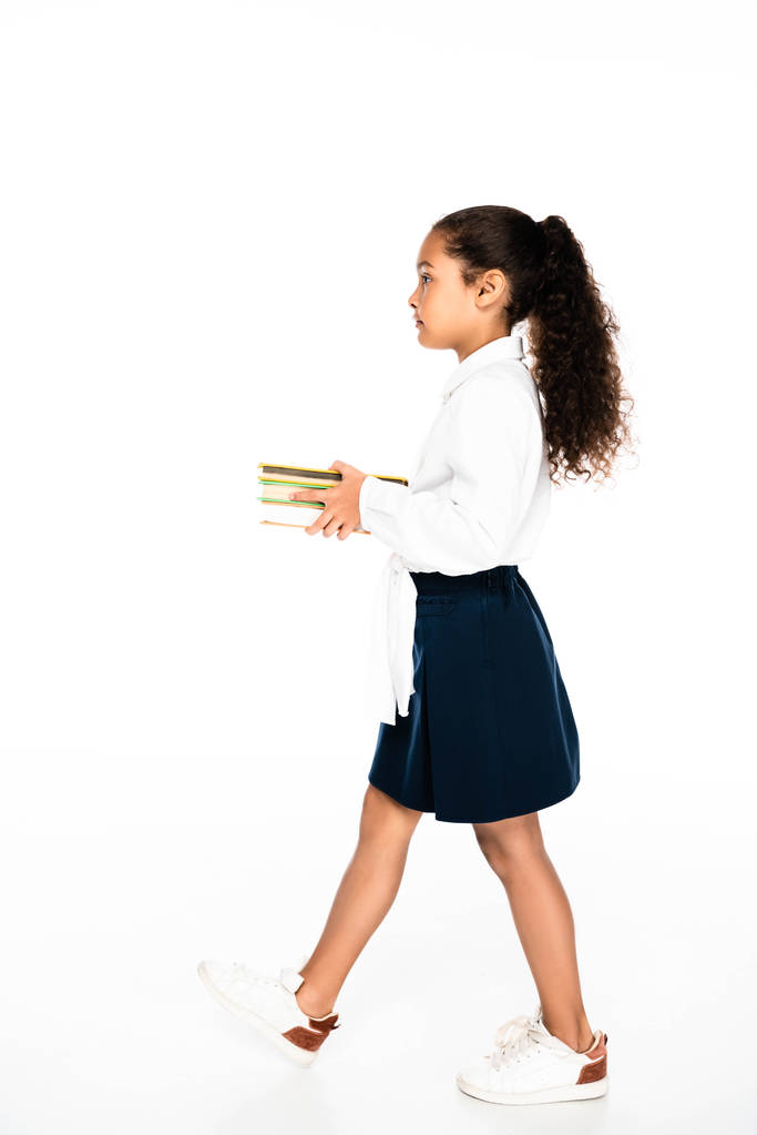 vue latérale de l'adorable écolière afro-américaine marchant tout en tenant des livres sur fond blanc
 - Photo, image
