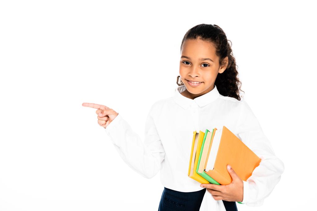 vrolijke African American schoolmeisje wijzend met vinger terwijl het houden van boeken geïsoleerd op wit - Foto, afbeelding