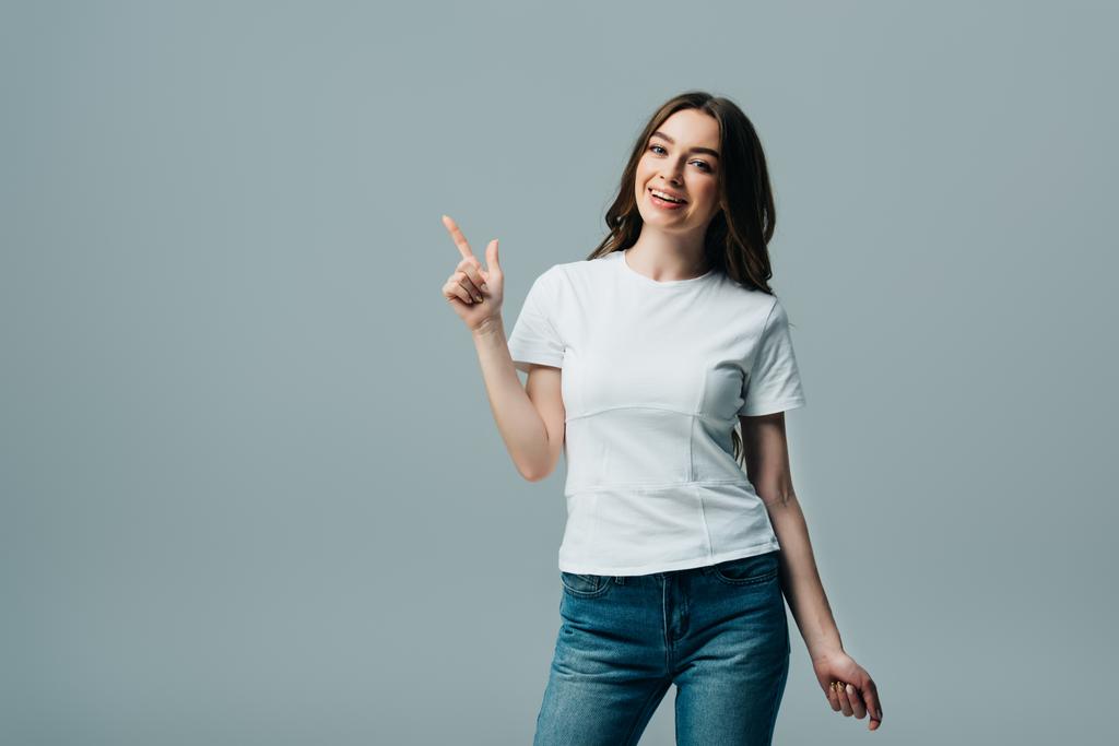 χαμογελαστή όμορφη κοπέλα με λευκό t-shirt που δείχνει με το δάχτυλο εκτός από το γκρι - Φωτογραφία, εικόνα