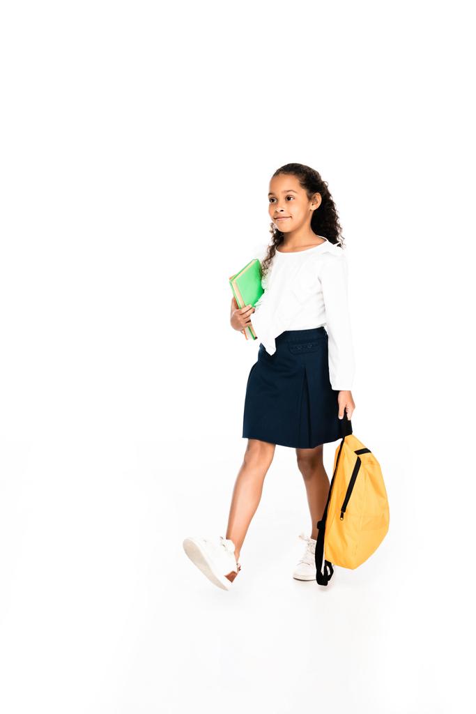 full length view of african american schoolgirl walking with backpack and book on white background - Photo, Image