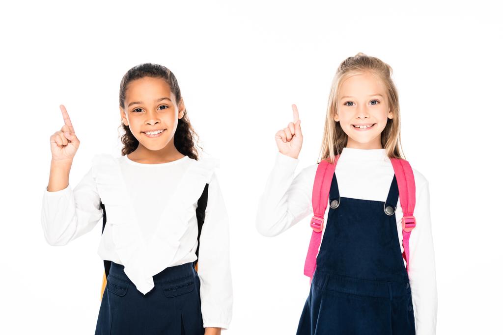dvě veselé multikulturní školačky zobrazující speciální gesta při úsměvu na kameře na bílém - Fotografie, Obrázek