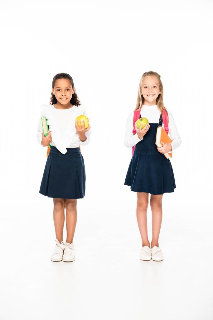volledige lengte weergave van twee glimlachende multiculturele schoolmeisjes houden van boeken en appels op witte achtergrond - Foto, afbeelding