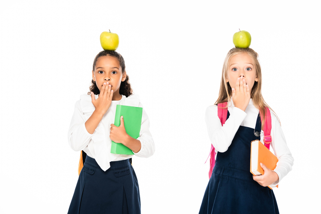 zwei schockierte multikulturelle Schulmädchen mit Äpfeln auf dem Kopf, die Münder bedecken, die Hände isoliert auf weiß - Foto, Bild