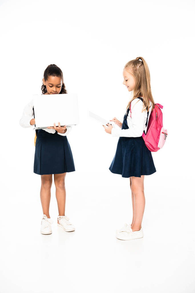 zobrazení celé délky dvou multikulturních školaček používajících přenosný počítač a digitální tablet na bílém pozadí - Fotografie, Obrázek